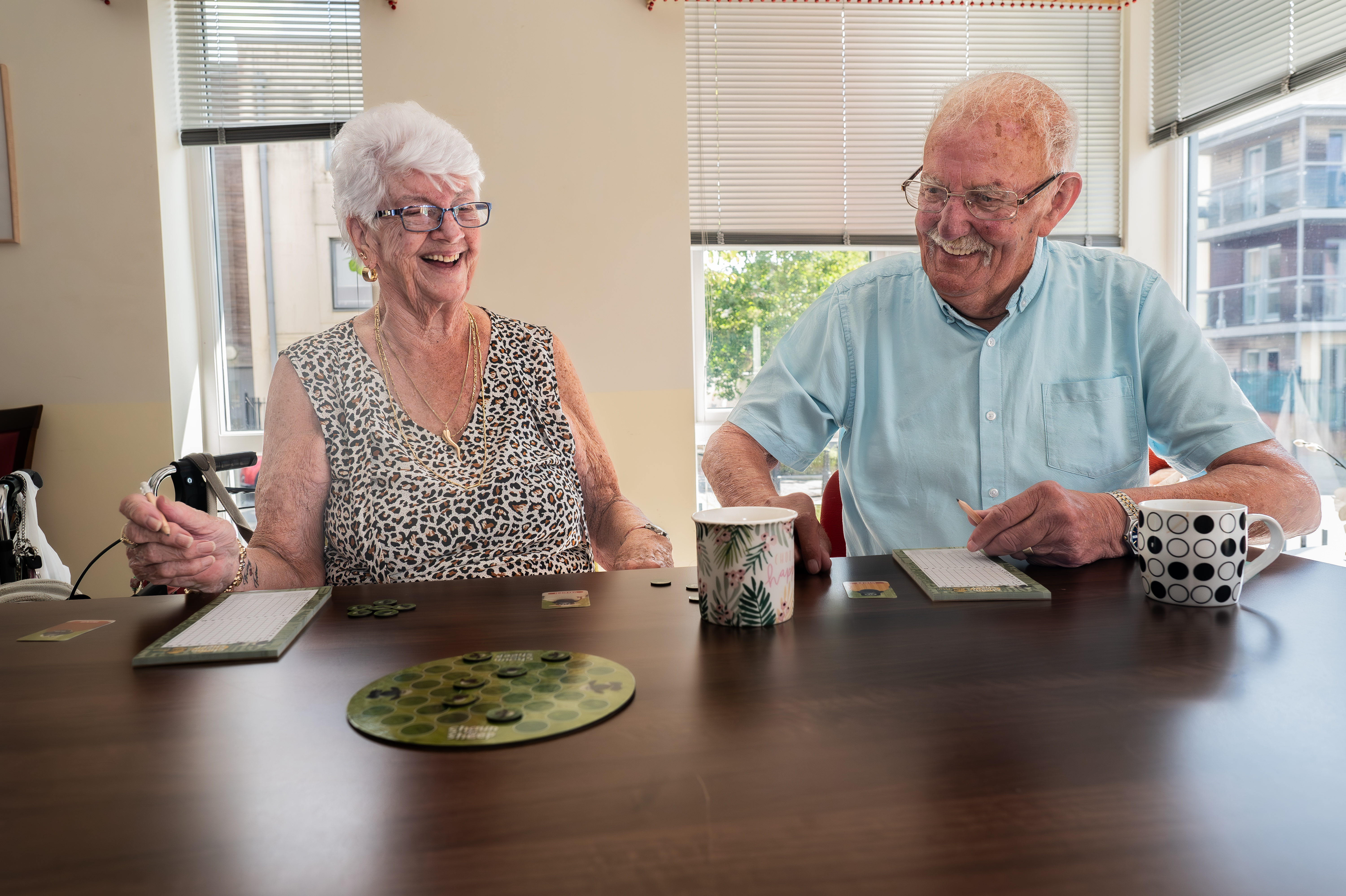 An elderly white couple sit at a table, smiling and laughing.