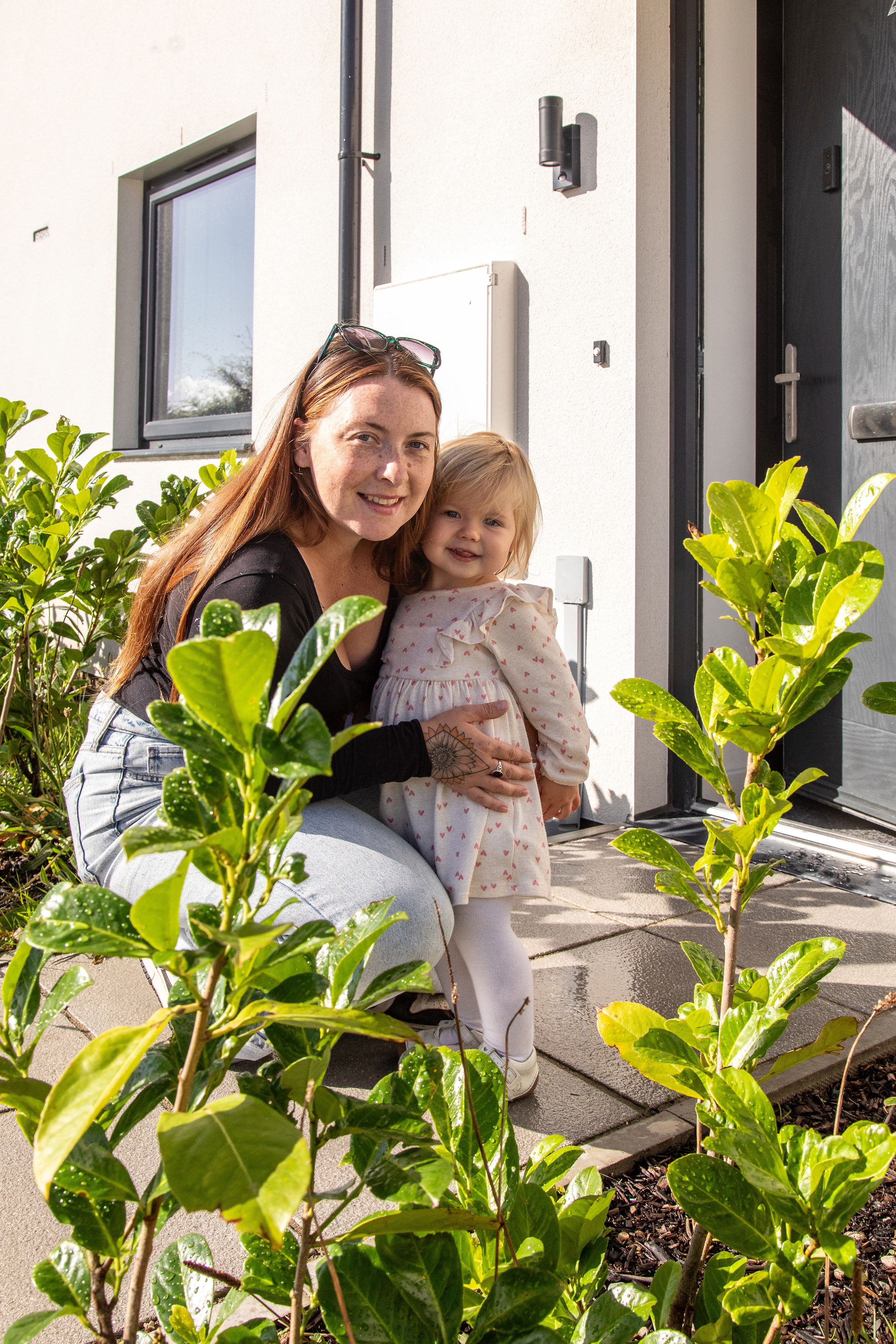 A parent and child, smiling and crouched in front of their bright white home exterior and grey front door. A small amount of foliage is in the foreground, and the photo is sunny and well lit. and the
